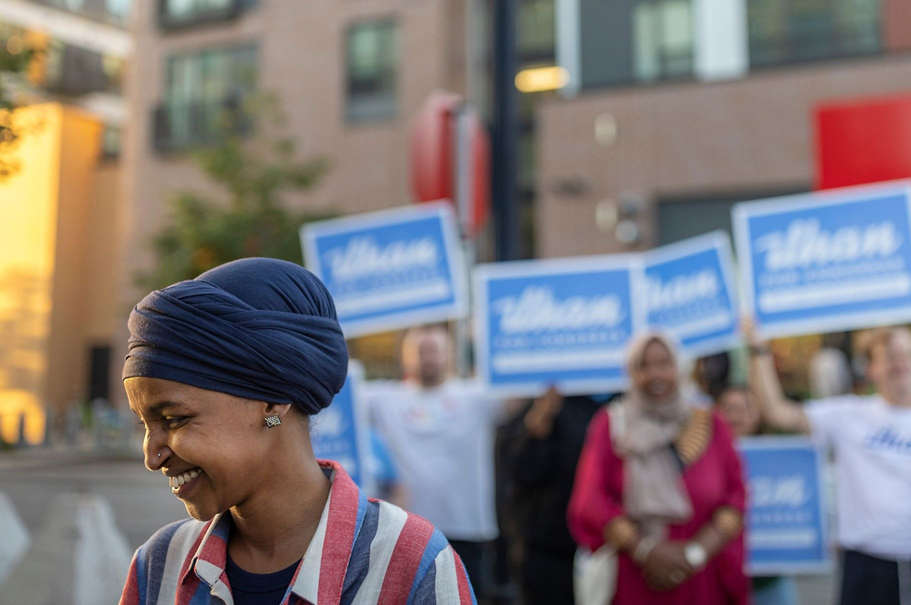 Omar looks down smiling, standing in front of a group of people holding "Ilhan for Congress" signs